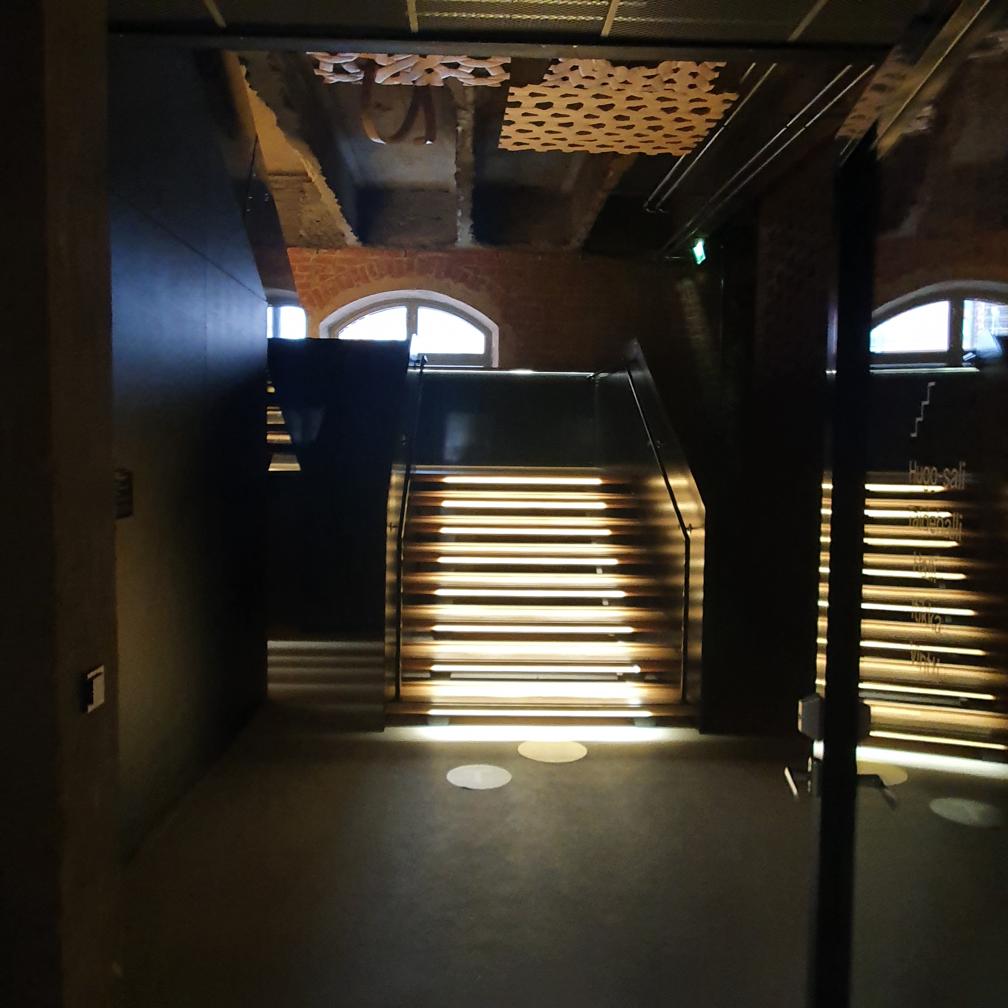 Three wooden sculptures resembling woven yarn patterns, suspended from ceiling above a wooden staircase. Photo taken from a distance of 5 metres.