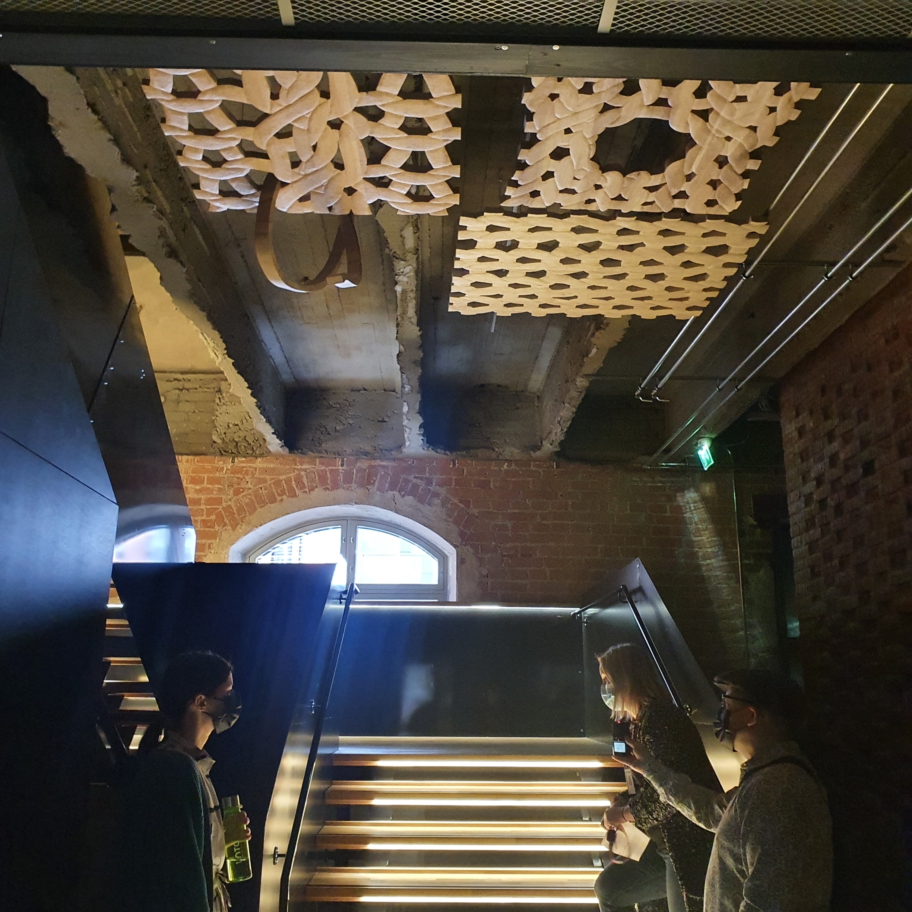 Three wooden sculptures resembling woven yarn patterns, suspended from ceiling above a wooden staircase