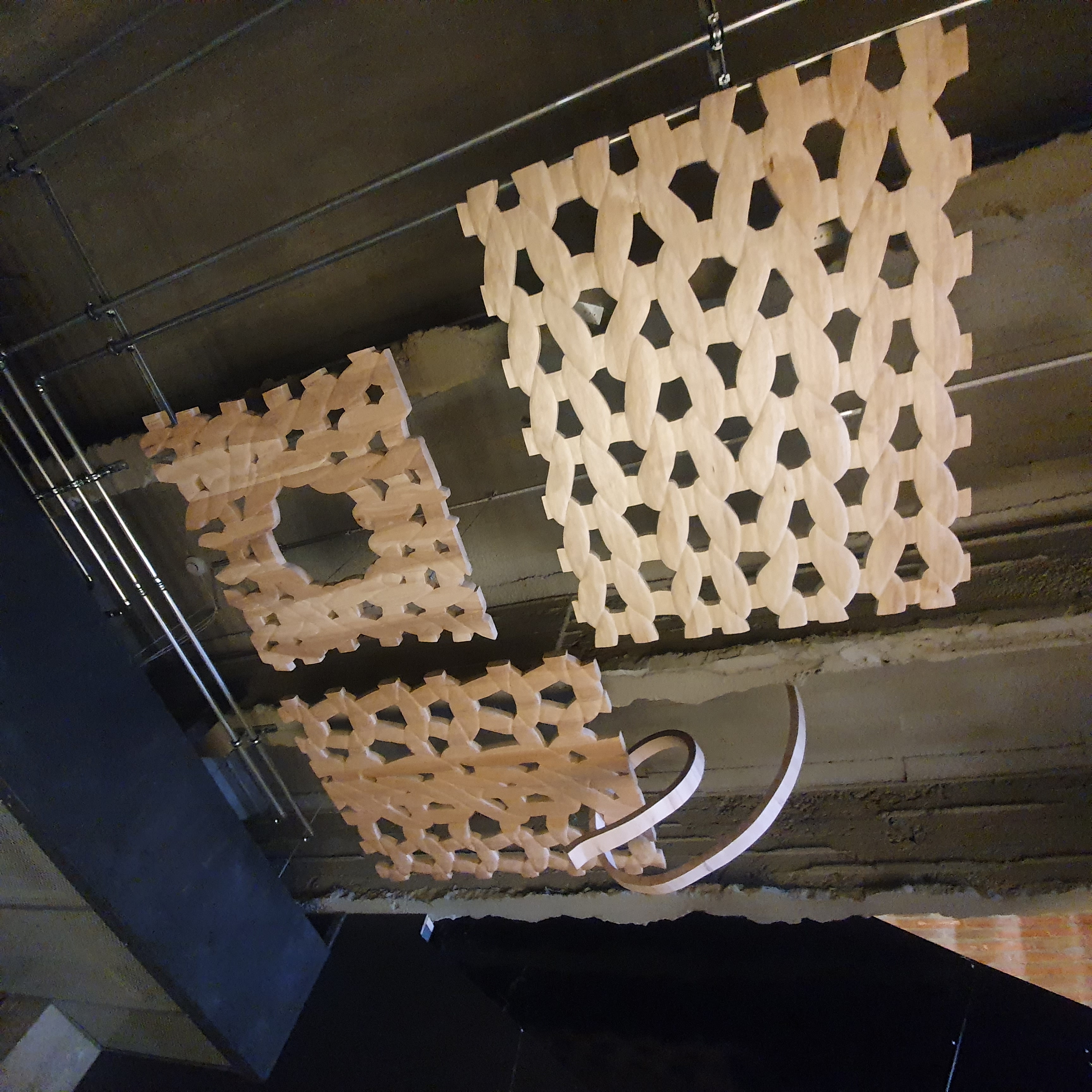 Three wooden sculptures resembling woven yarn patterns, suspended from a concrete ceiling
