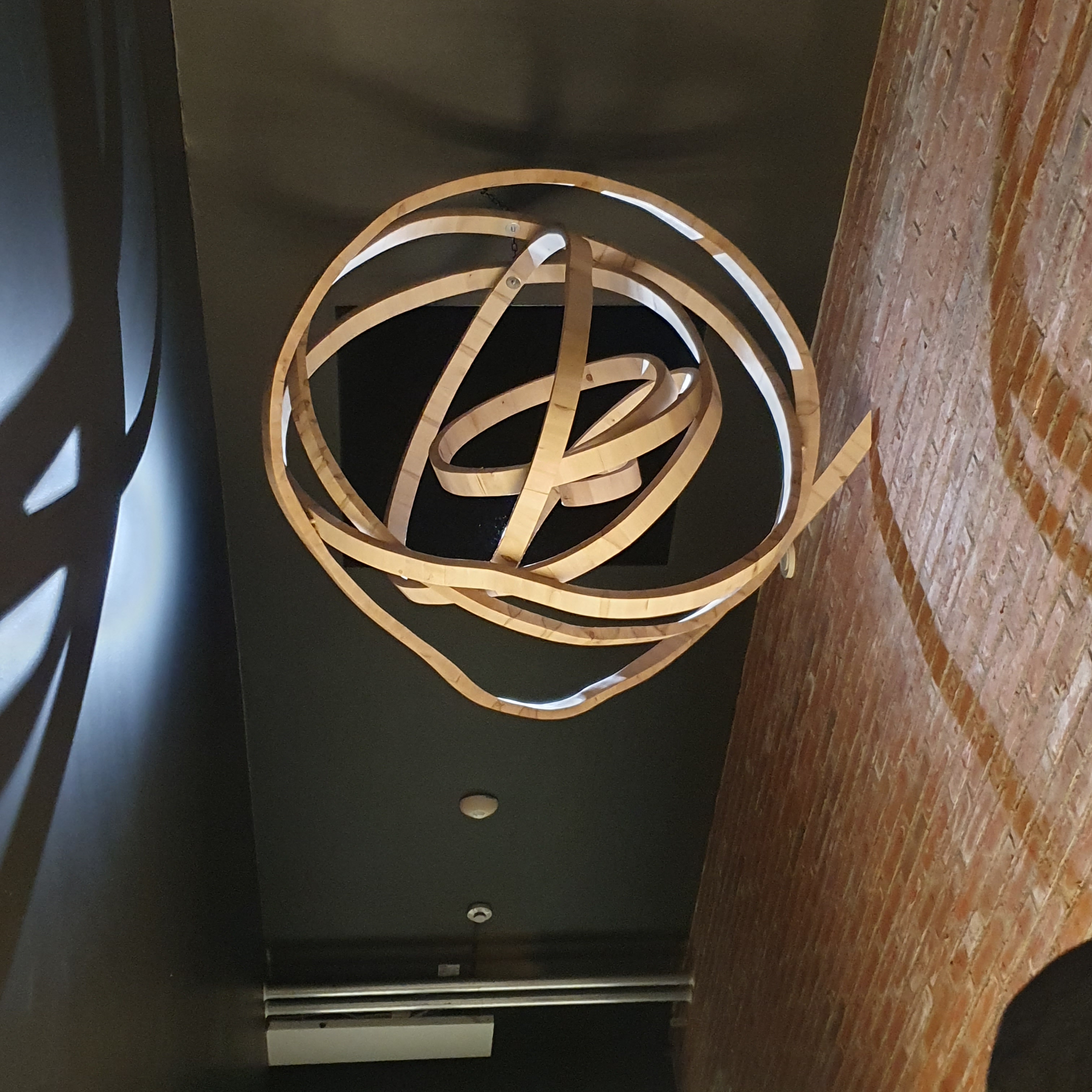 Wooden sculpture resembling a ball of yarn, suspended from the ceiling alongside a red brick wall