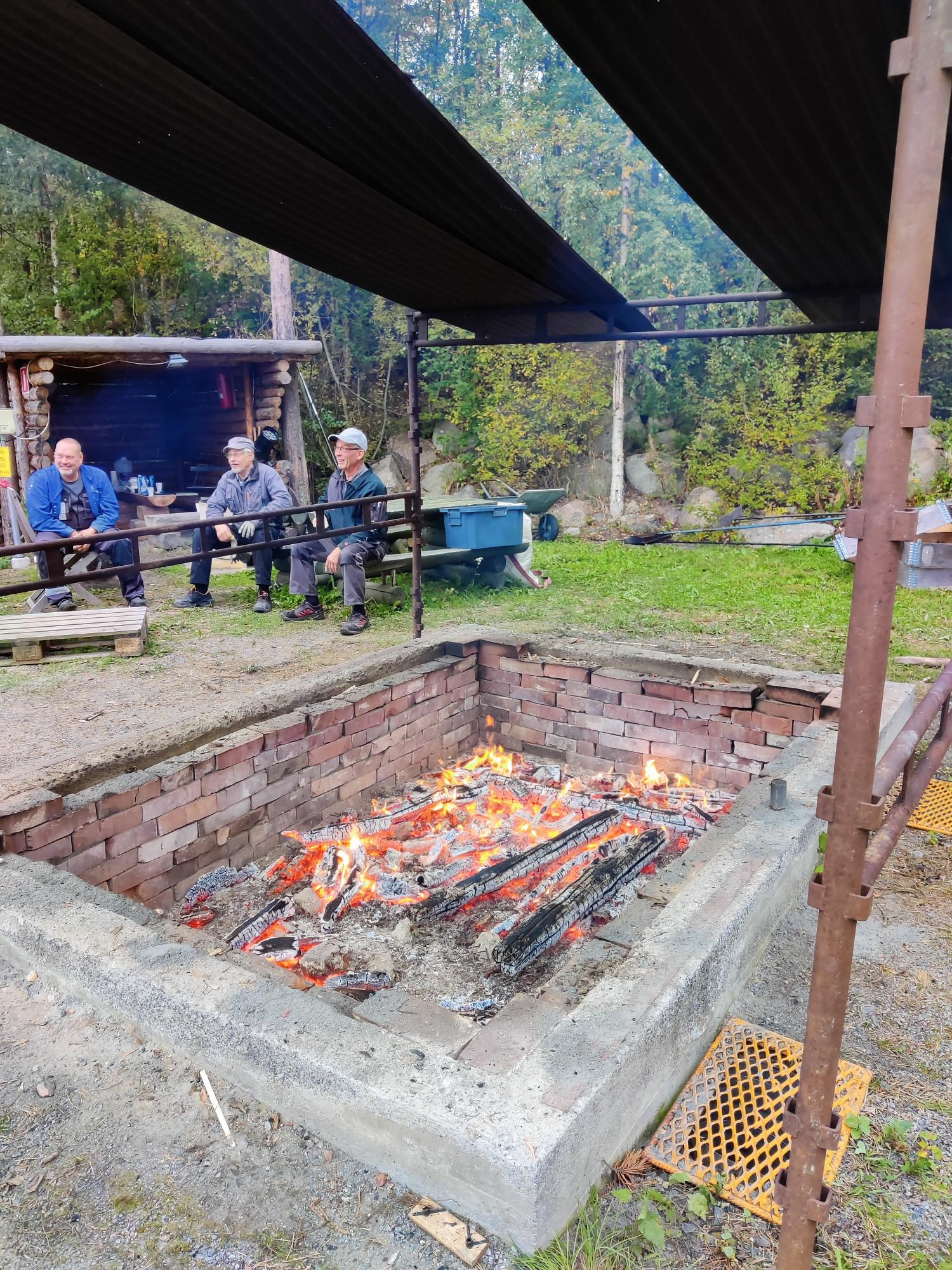 Men sitting at a table next to a large, open fire-pit for grilling food