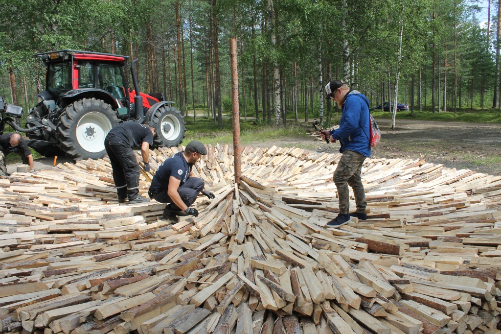 Two men placing small wooden logs to build a tar pit, with a bearded Australian filming them with a phone.