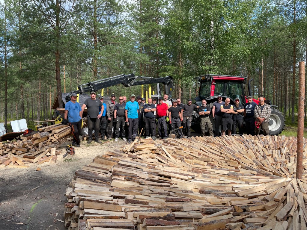 Lions club team. Group of men standing in a line around a woodpile building a tar pit.