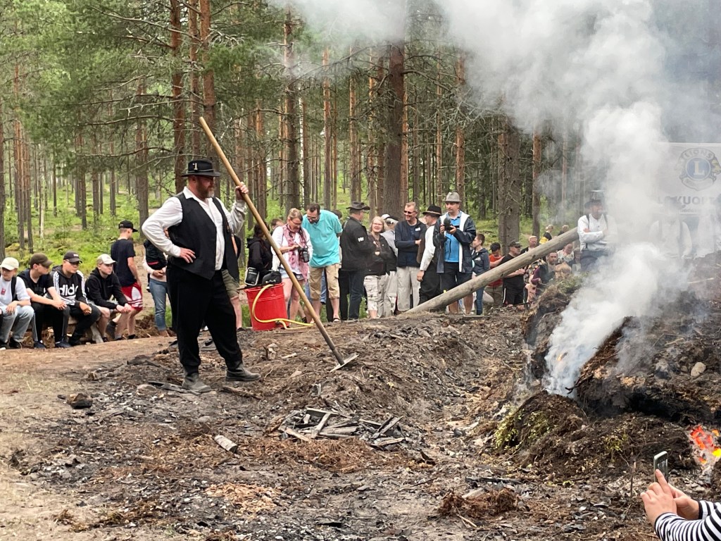 Man in traditional costume tends to the fire with a metal hook on a long wooden pole.