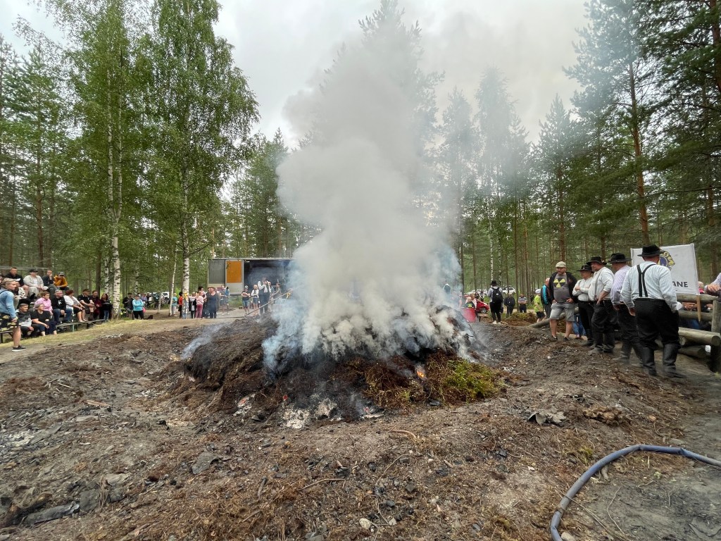 Smouldering fire pit with billowing white smoke and people sitting around on bleachers/stands.